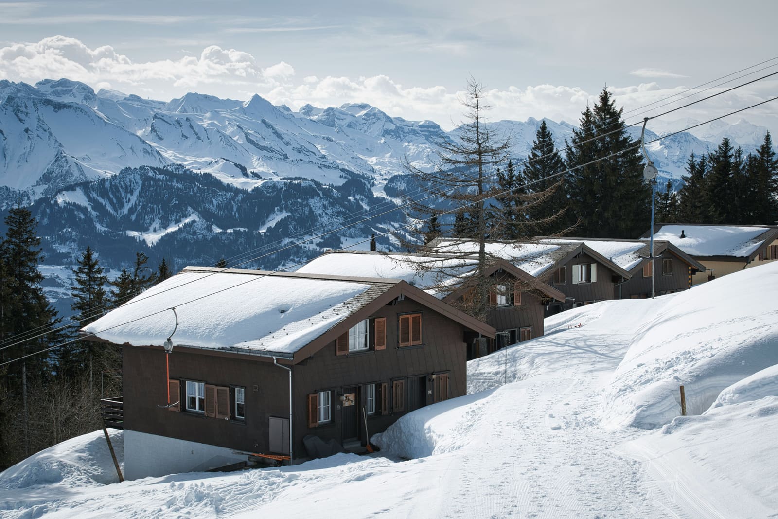 Verschneite Holzchalets an einem Dorfweg vor einem weiten Alpenpanorama