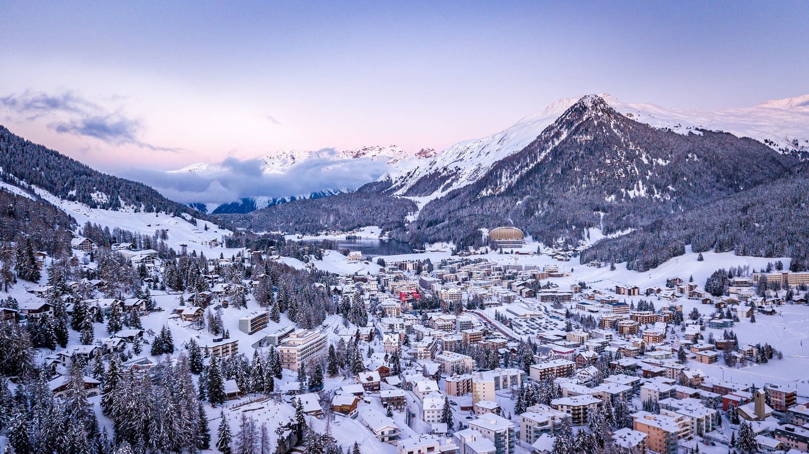 Luftaufnahme eines grossen verschneiten Bergorts mit See und Gipfeln in der Morgendämmerung