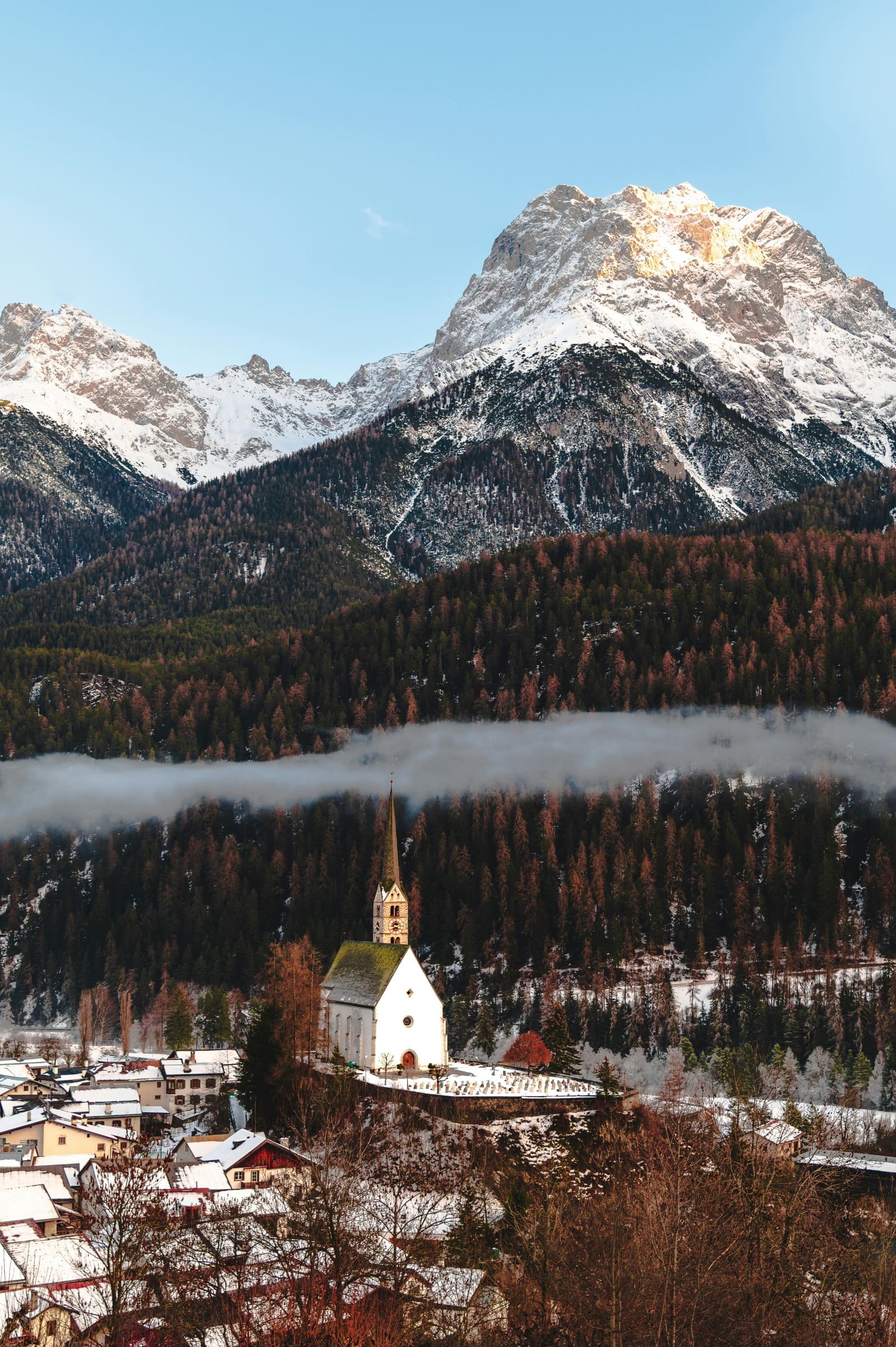 Weisse Kirche über einem Dorf, dahinter Nebelband und ein schneebedeckter Gipfel