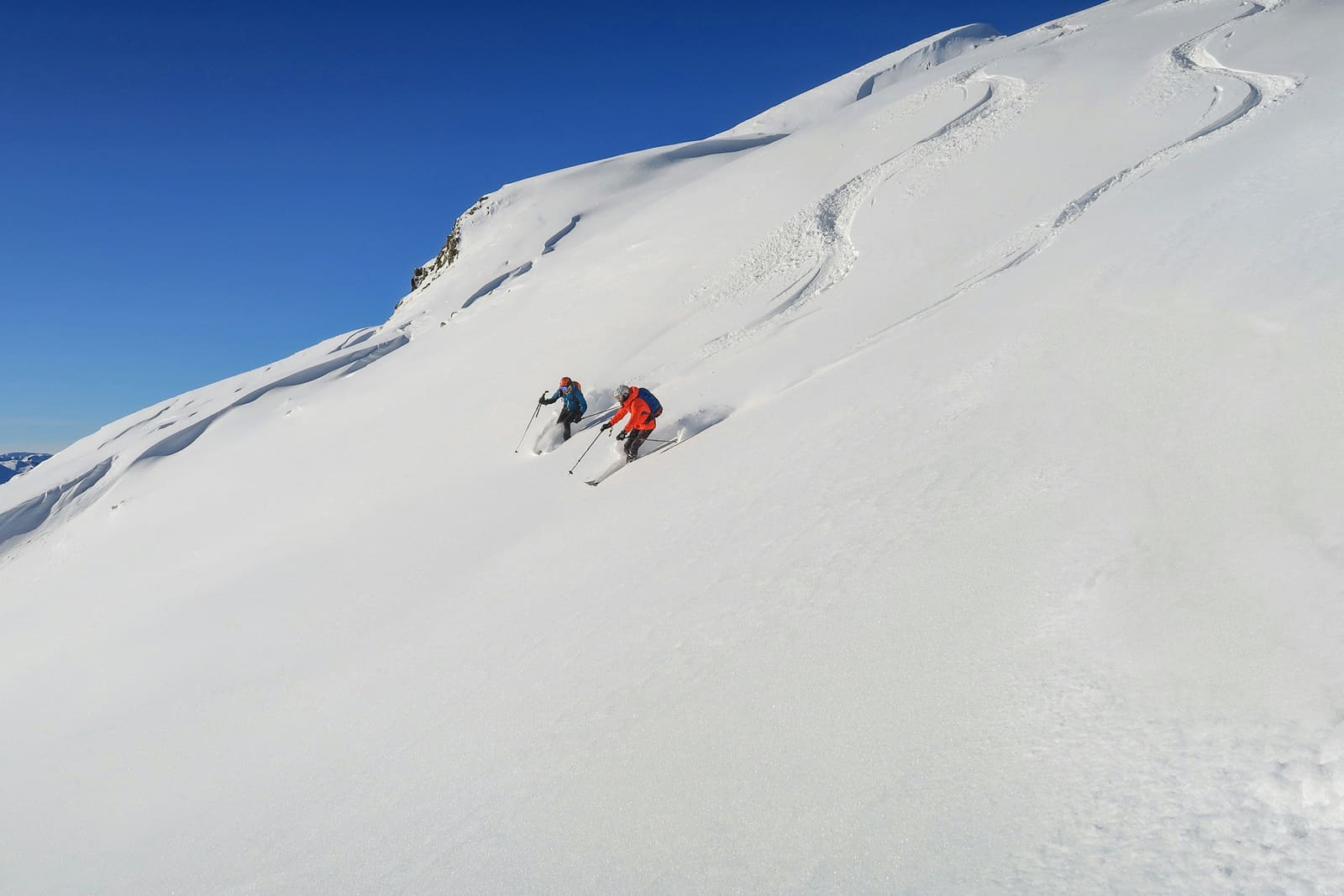Zwei Freerider ziehen Spuren in einen unberührten Pulverschneehang unter blauem Himmel