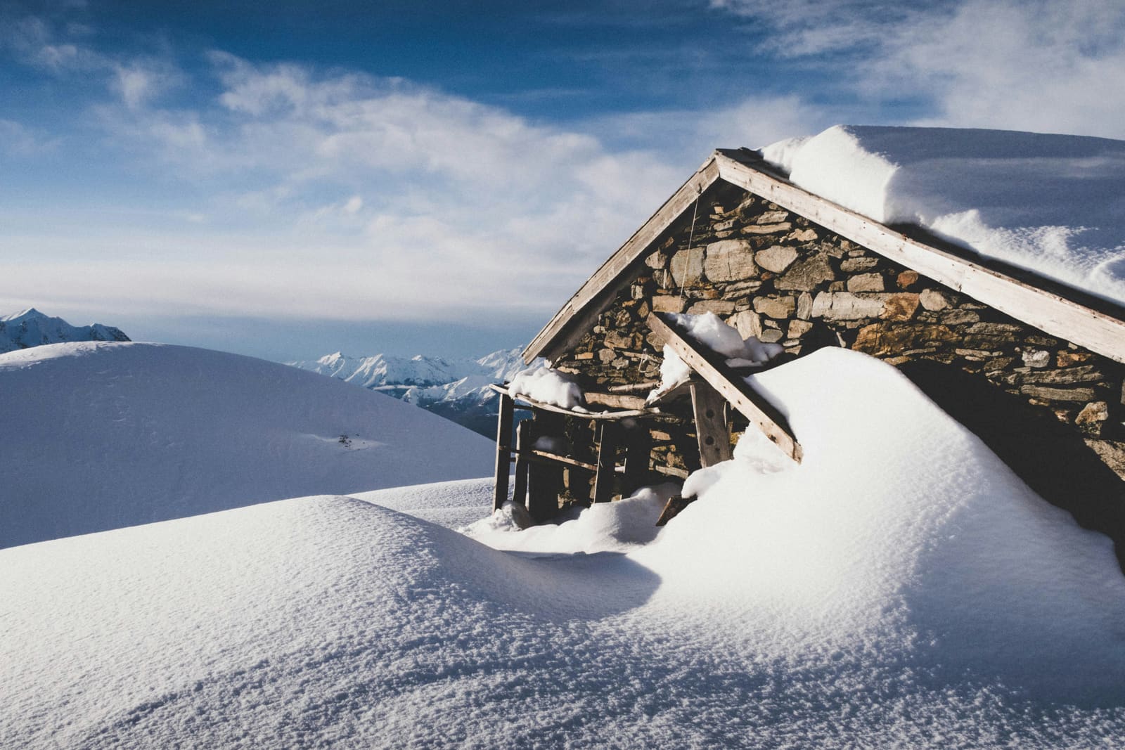 Tief verschneite Steinhütte auf einer Anhöhe mit Bergpanorama im Hintergrund