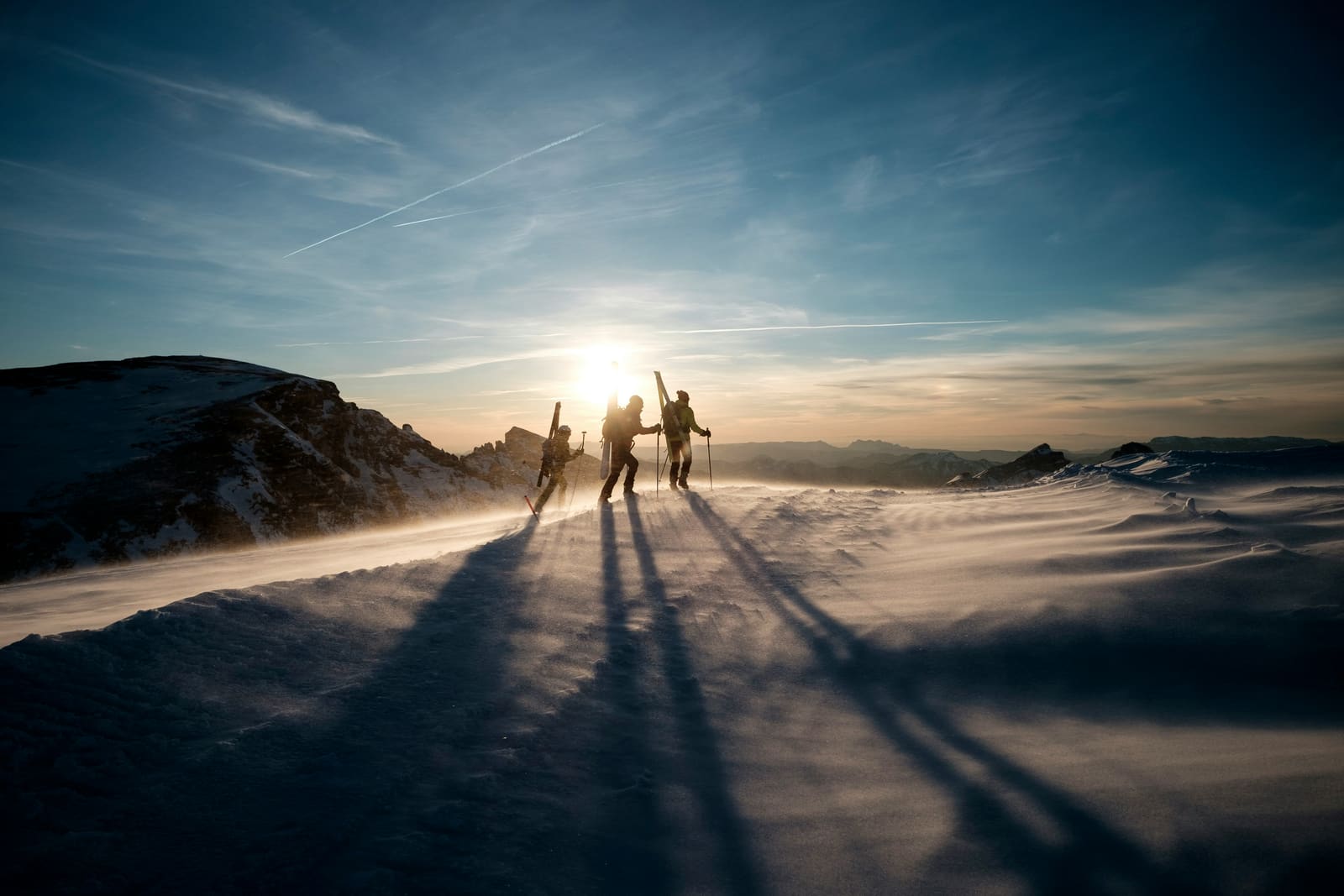 Skitourengruppe mit Ski am Rucksack im Gegenlicht auf windigem Grat bei Sonnenuntergang